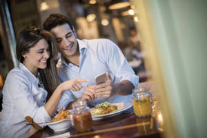Man and woman sitting at table with food looking at a cell phone.
